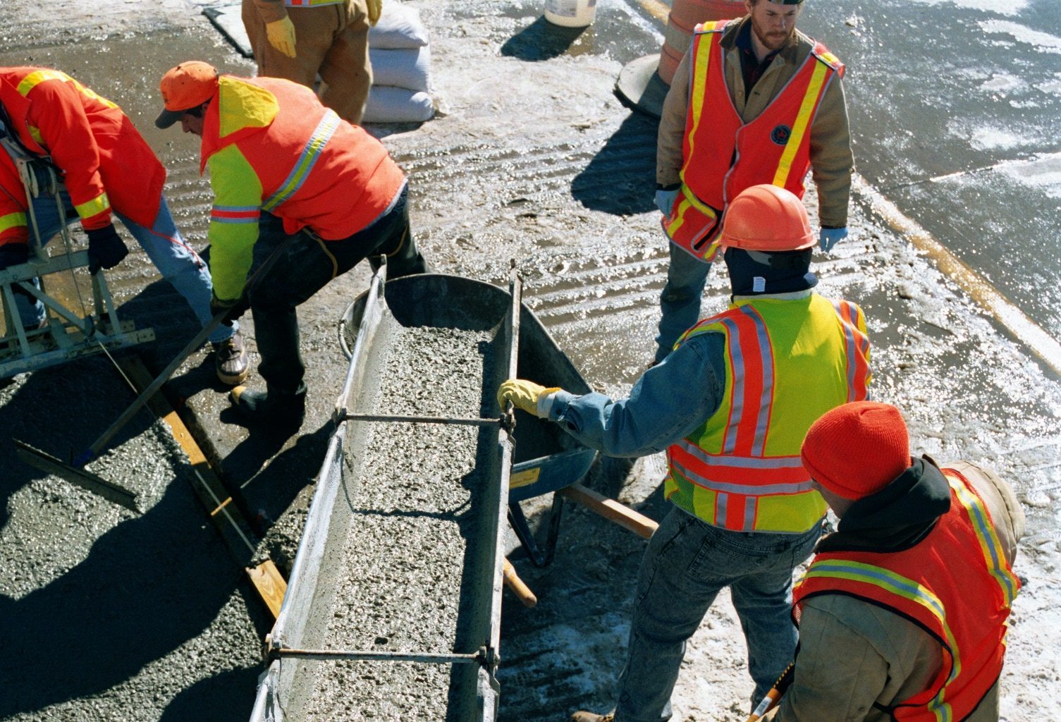 Several construction workers in hard hats and safety vests are pouring concrete at a worksite to create sustainable architecture. 